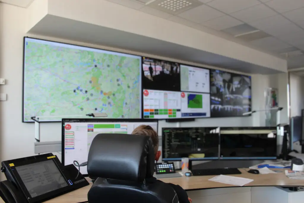 Operator zit voor een workstation. Aan de muur toont een videowall een verzameling van kaarten en andere informatie